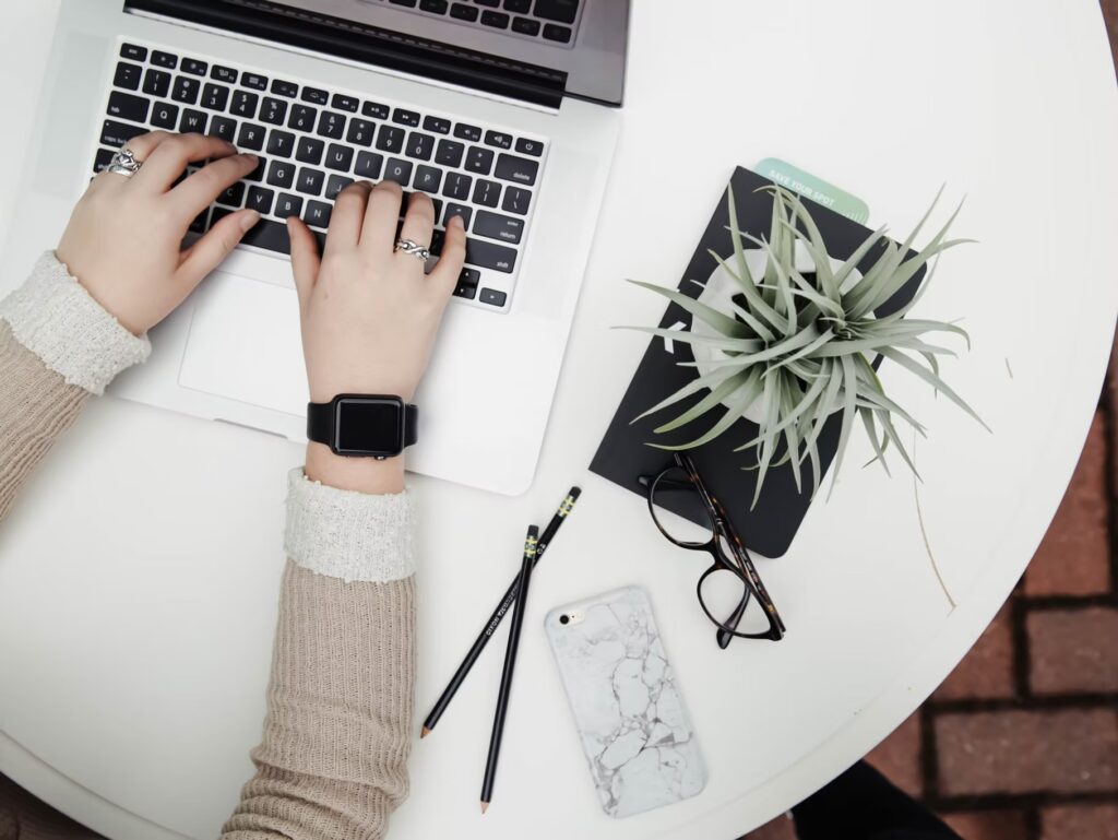 Person typing on a laptop at a round white table with a plant, notebook, glasses, pencils, and a marble-patterned phone nearby.