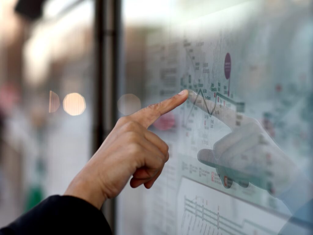 A person points at a location on a glass-covered map or transit schedule at a public transportation stop.