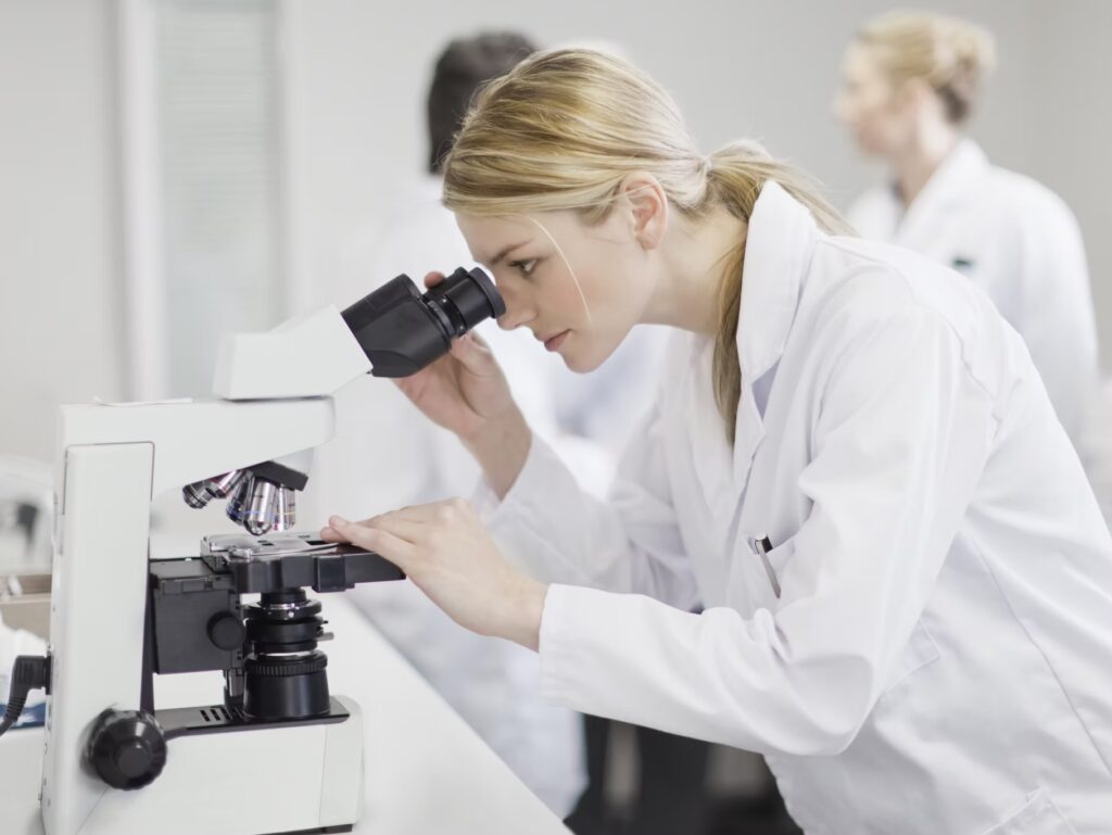 A woman in a white lab coat looks through a microscope in a laboratory, with two other people in lab coats blurred in the background.