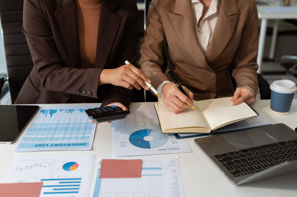 Two people in business attire review financial charts and documents at a desk, with a laptop, calculator, coffee cup, and open notebook.