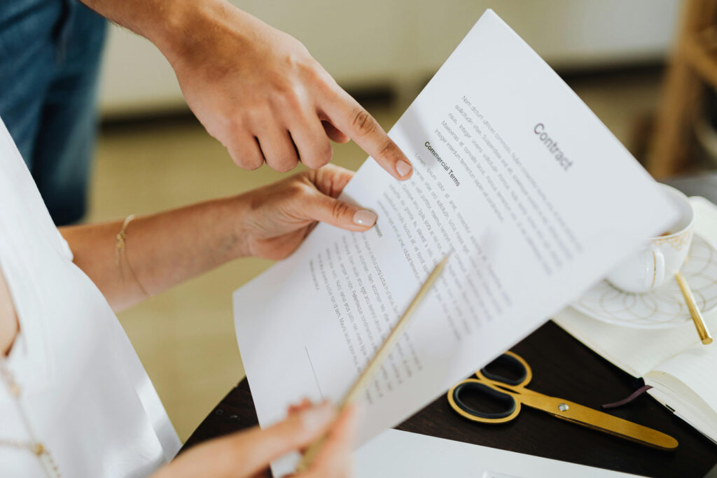 Two people review and point at a printed contract document on a desk with a pair of glasses, a pencil, and a cup nearby.