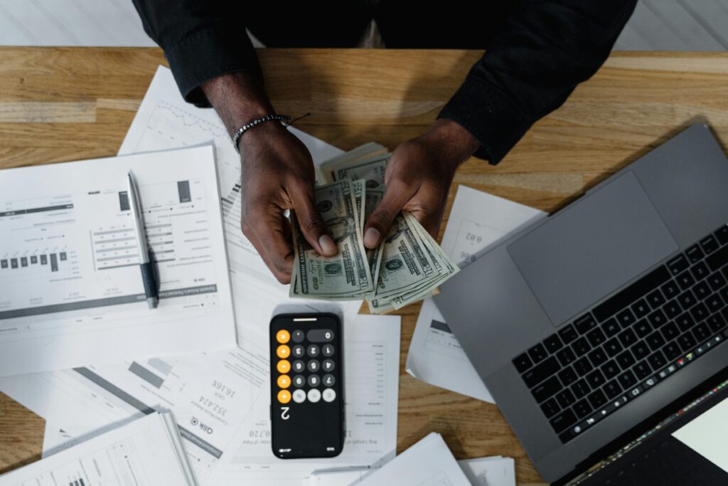 Person counting US dollar bills over financial documents with a calculator, pen, and laptop on a wooden desk.