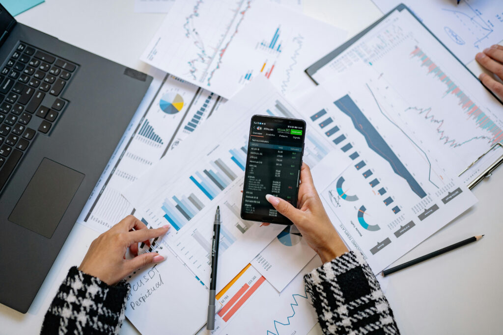 Person analyzing financial charts and graphs on paper at a desk, holding a smartphone displaying stock market data, with a laptop and pens nearby.
