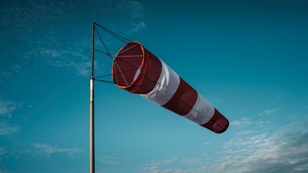 A red and white striped windsock attached to a tall pole stands against a clear blue sky with a few scattered clouds.
