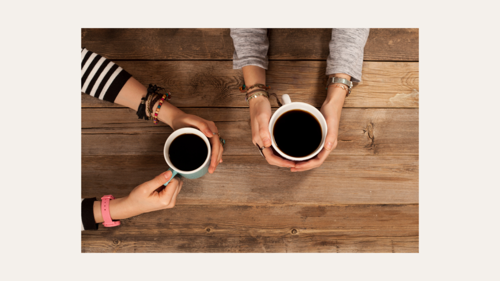 Two people sit at a wooden table holding mugs of black coffee, with only their arms and hands visible.